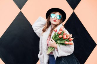 Happy emotional fashion woman in heart-shaped sunglasses holding bouquet on peach color background